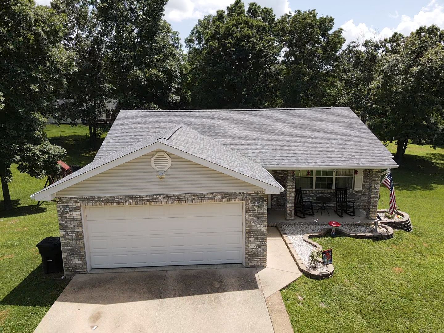House with gray roof, tan siding, and stone accents. Features a garage, porch, and American flag in a grassy yard.