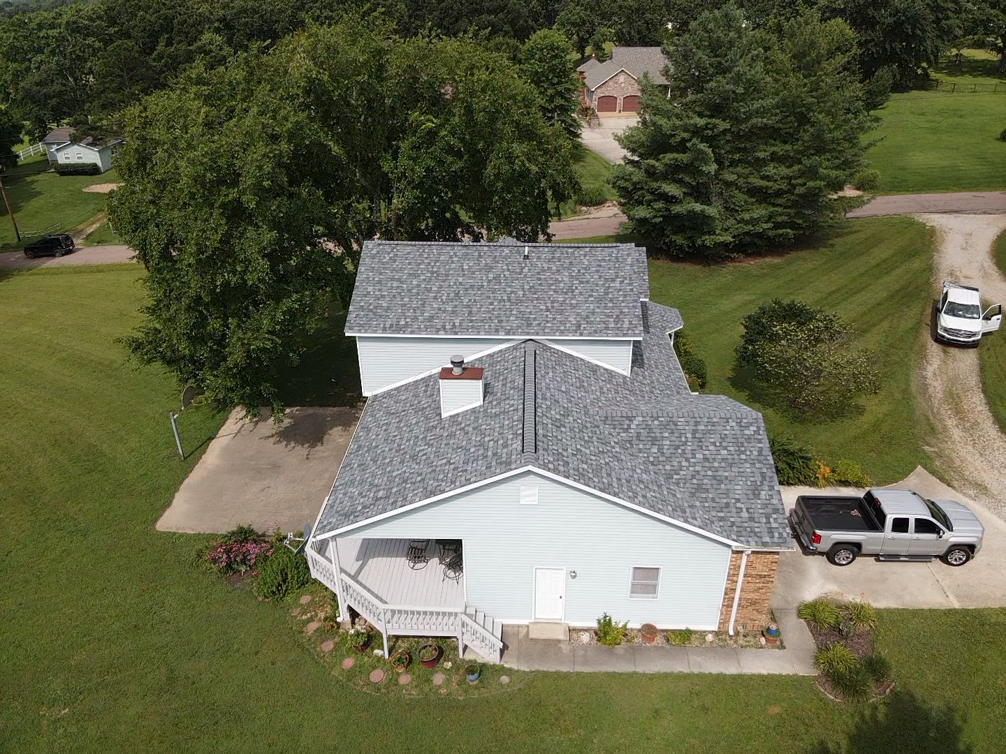 Aerial view of a light blue house with a gray roof, surrounded by green grass and trees. A truck and SUV are parked nearby.