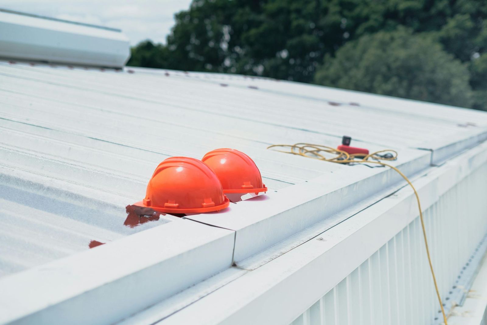 Two orange hard hats on a white metal roof with safety rope and green trees in the background.